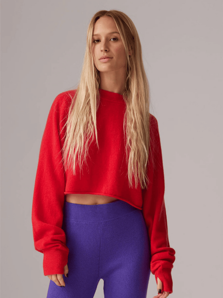 Eine blonde Frau trägt einen roten, cropped Pullover mit überdimensionalen Ärmeln. Sie steht vor einem neutralen Hintergrund und trägt lila, figurbetonte Hosen.